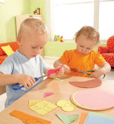 Zwei kleine Kinder sitzen an einem Tisch und schneiden mit einer Schere Formen aus farbigem Papier aus. Der Tisch ist mit Papier und Bastelmaterial bedeckt. Der Hintergrund zeigt ein helles, aufgeräumtes Zimmer mit Spielzeug und Möbeln.