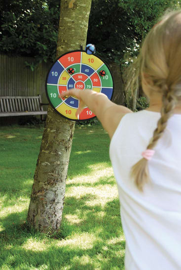 Ein Kind mit blondem Zopf und weißem Hemd wirft einen Klettball auf eine bunte Dartscheibe, die an einem Baum in einem sonnigen, grasbewachsenen Hinterhof befestigt ist. Im Hintergrund ist eine Holzbank zu sehen.
