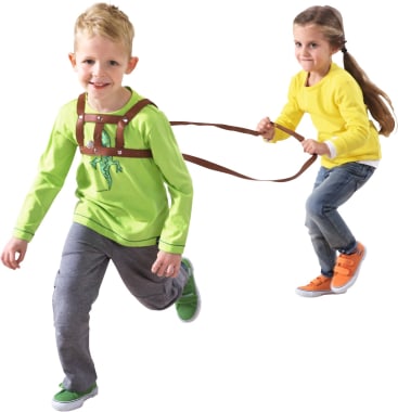 Zwei Kinder spielen. Ein Kind in grünem Hemd und grauer Hose läuft voraus, während ein anderes Kind in gelbem Hemd und Jeans sich an einem Geschirr und einer Leine festhält, die am ersten Kind befestigt sind.
