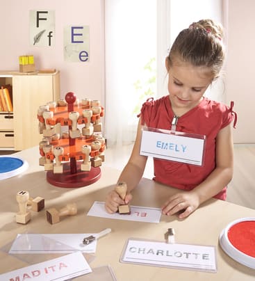 Ein junges Mädchen in einem Klassenzimmer benutzt einen Stempel, um eine Namenskarte mit der Aufschrift EMELY zu markieren. Sie trägt ein Namensschild, und auf dem Tisch liegen weitere Namenskarten, Stempel und Stempelkissen. An der Wand hängen Alphabet-Poster.