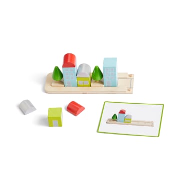 Ein Holzspielzeug-Stadtbausatz mit farbigen Blöcken, die Gebäude und Bäume auf einer rechteckigen Basis bilden, neben losen Blöcken und einer Karte, die ein Bild des fertigen Bauwerks zeigt.