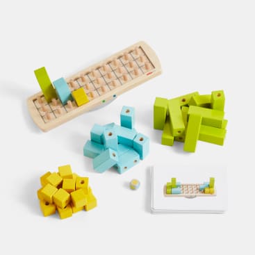 Ein hölzernes Spielset mit einem rechteckigen Spielbrett, Stapeln von grünen, blauen und gelben Blöcken, einem kleinen Würfel und einem Satz illustrierter Karten, die auf einer weißen Fläche angeordnet sind.