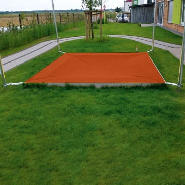 Ein rechteckiges orangefarbenes Sonnensegel ist über einer gepflasterten Fläche in einem grasbewachsenen Hinterhof mit einem Gehweg, jungen Bäumen und einem Gebäude auf der rechten Seite installiert.