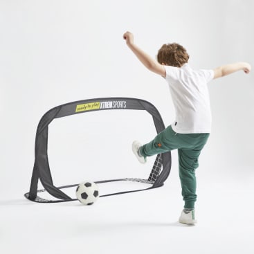 Ein Kind in weißem Hemd und grüner Hose kickt einen Fußball in Richtung eines kleinen schwarz-weißen XTREM SPORTS-Tornetzes auf einem schlichten weißen Hintergrund.