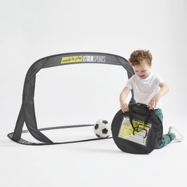 Ein kleiner Junge kniet neben einem kleinen Fußballtor mit einem Fußball und hält eine schwarze Tragetasche mit einem Produktetikett. Auf dem Tor ist "Ready to play" von XTREM SPORTS aufgedruckt. Der Hintergrund ist weiß.