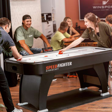 Zwei Personen spielen Airhockey an einem schwarz-weißen Tisch mit der Aufschrift Speed Fighter Air Hockey, während mehrere andere Personen in einer ungezwungenen Innenumgebung sitzen und zusehen.