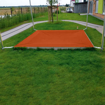 Ein rechteckiges orangefarbenes Schattentuch ist horizontal zwischen vier Metallstangen auf einem grasbewachsenen Rasen mit gepflasterten Gehwegen und einem kleinen Baum im Hintergrund aufgespannt.