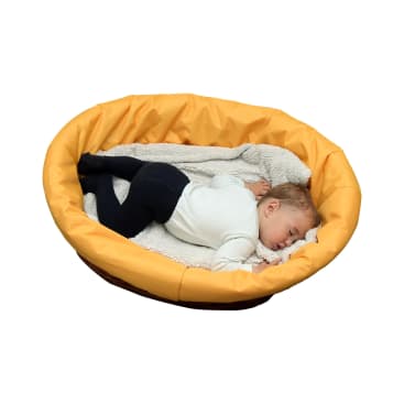 Ein kleines Kind schläft auf der Seite in einem runden, gelben, gepolsterten Haustierbett, das mit einer weichen, hellen Decke ausgekleidet ist. Der Hintergrund ist einfarbig weiß.
