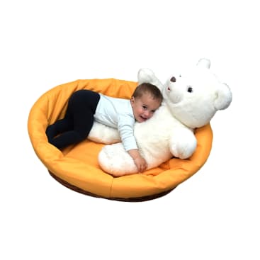 Ein kleines Kind in einem hellblauen Hemd und einer dunklen Hose liegt auf einem großen weißen Teddybären in einem orangefarbenen, gepolsterten runden Bett, lächelt und umarmt das Spielzeug. Der Hintergrund ist weiß.