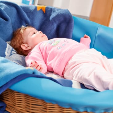Ein Baby liegt auf dem Rücken in einem Korbbett mit blauem gepolstertem Rand und einer blauen Decke. Das Baby ist rosa gekleidet und blickt nach oben.