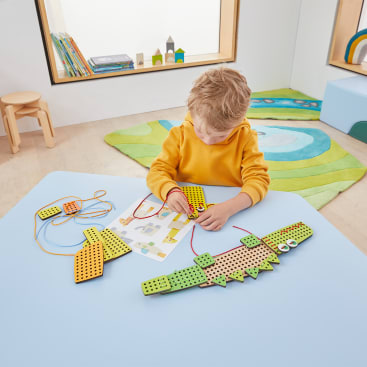 Ein Kind in einem gelben Pullover sitzt an einem blauen Tisch und baut eine krokodilförmige Bastelarbeit mit gelochten Teilen und Schnüren zusammen. Anleitungen und zusätzliche Teile liegen auf dem Tisch in einem hellen, modernen Spielzimmer.