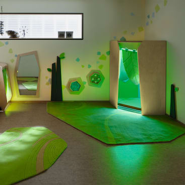 Ein modernes Spielzimmer mit grünen Teppichen, geometrischen Wanddekorationen, einem rechteckigen Spiegel und einer hölzernen Spielstruktur mit einem beleuchteten Eingang und grünem Licht, das von innen leuchtet. Durch ein hohes Fenster fällt natürliches Licht ein.