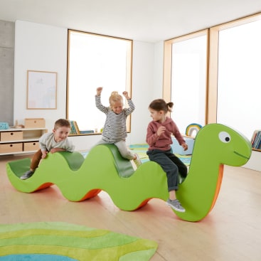 Drei kleine Kinder spielen in einem hellen Klassenzimmer mit Holzfußboden und großen Fenstern auf einer großen, grünen, raupenförmigen Softplay-Struktur.