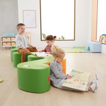 Drei kleine Kinder sitzen auf grünen, schlangenförmigen Kissen in einem hellen Klassenzimmer und lesen jeweils ein Buch. Der Raum ist mit Holzböden, Regalen und Kinderzeichnungen an den Wänden ausgestattet.