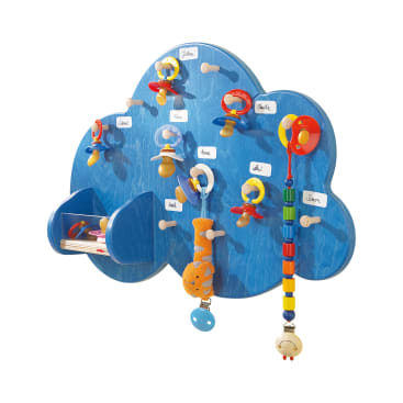 Ein blauer, wolkenförmiger Wandhalter mit beschrifteten Haken für bunte Schnuller, eine Rassel und ein Perlenspielzeug. In einem kleinen Regal stehen mehrere Kinderbücher. Die Etiketten sind mit handgeschriebenen Namen versehen.