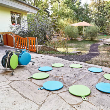Außenbereich mit Steinpflaster, verstreuten blauen und grünen runden Sitzkissen, einem Wagen mit weiteren Kissen, einem orangefarbenen Zaun, einem gelben Gebäude und einem Garten mit Bäumen, Sträuchern und einem großen beigen Sonnenschirm im Hintergrund.