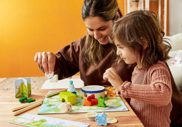 Ein Erwachsener und ein Kind sitzen an einem Tisch und spielen ein Brettspiel mit Tierfiguren und bunten Spielsteinen. Im Hintergrund sind eine orangefarbene Wand und eine beigefarbene Couch zu sehen.