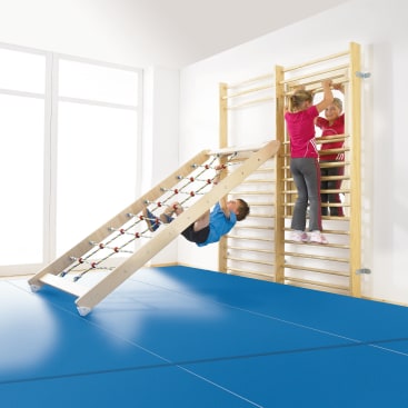 Zwei Kinder klettern an einer hölzernen Sprossenwand in einem hellen Raum mit großen Fenstern, während ein anderes Kind an einer Seilrampe hängt, die an den Sprossen befestigt ist. Blaue, gepolsterte Matten bedecken den Boden zur Sicherheit.