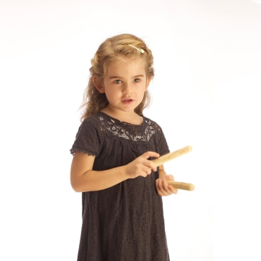 Ein junges Mädchen mit hellbraunem Haar, das ein dunkles Spitzenkleid trägt, hält zwei hölzerne Rhythmus-Sticks vor einem schlichten weißen Hintergrund.