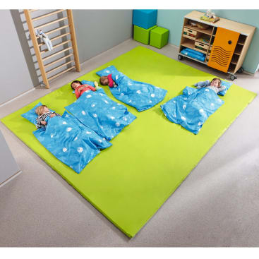 Vier kleine Kinder liegen auf einer großen grünen Matte in einem Spielzimmer, jedes mit einer blauen Decke mit weißen Wolken bedeckt; im Hintergrund stehen Regale und Turngeräte.
