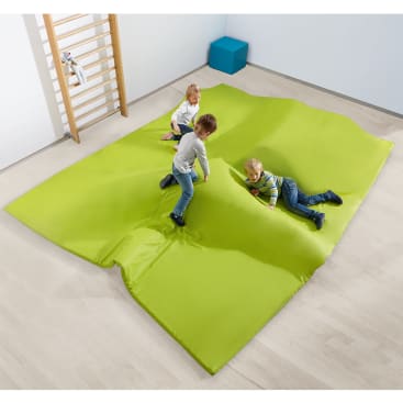 Drei kleine Kinder spielen auf einer großen, grün gepolsterten Matte in einem hellen Raum mit Holzfußboden. Im Hintergrund sind ein hölzernes Klettergerüst und ein blauer Kasten zu sehen.