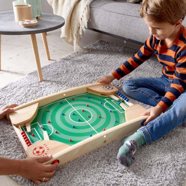Ein Kind sitzt auf einem grauen Teppich und spielt mit einer anderen Person ein Tischfußballspiel. Das Spiel hat ein grünes Feld, kleine farbige Pflöcke und Holzschläger. Im Hintergrund sind ein Tisch und eine Couch zu sehen.