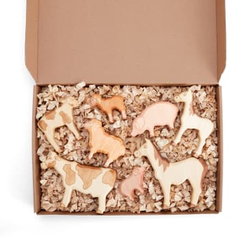 Eine mit Holzspänen gefüllte Pappschachtel enthält sieben Holztierfiguren, darunter eine Kuh, ein Pferd, eine Ziege, ein Schwein, eine Giraffe, einen Hund und ein kleines Ferkel. Die Schachtel ist geöffnet und die Figuren sind fein säuberlich angeordnet.
