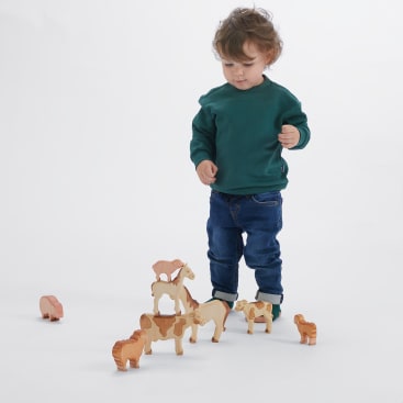 Ein kleines Kind in grünem Sweatshirt und blauer Jeans steht vor einem weißen Hintergrund und betrachtet Tierfiguren aus Holz, die in einer Gruppe auf dem Boden angeordnet sind.