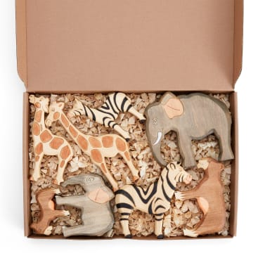 Eine offene, mit Holzspänen gefüllte Pappschachtel enthält sechs hölzerne Tierfiguren, darunter eine Giraffe, ein Elefant, zwei Zebras, ein Löwe und ein Affe.