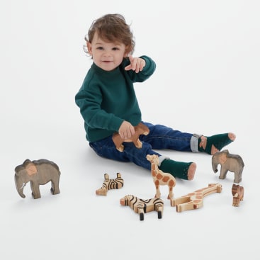 Ein kleines Kind in grünem Pullover und Jeans sitzt auf dem Boden, hält einen hölzernen Löwen in der Hand und zeigt nach vorne. Mehrere hölzerne Tierfiguren, darunter ein Elefant, eine Giraffe und ein Zebra, stehen verstreut herum.
