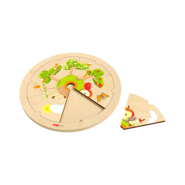 Ein rundes Holzpuzzle mit einem farbenfrohen Baum- und Tiermotiv, dem ein keilförmiges Teil fehlt, das neben das Puzzle gelegt wird. Das Puzzle ist von HABA Pro.
