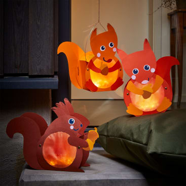 Drei Papierlaternen in Form von Eichhörnchen mit leuchtenden orangefarbenen Bäuchen sind ausgestellt; zwei hängen, und eine steht auf einem Kissen im Freien in der Nähe einer Gebäudewand.