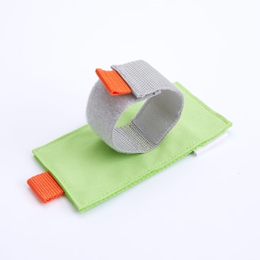 Ein graues Stoffarmband mit einer orangefarbenen Zuglasche liegt auf einer rechteckigen, hellgrünen Polstermatte, die ebenfalls eine orangefarbene Lasche hat, vor einem schlichten weißen Hintergrund.