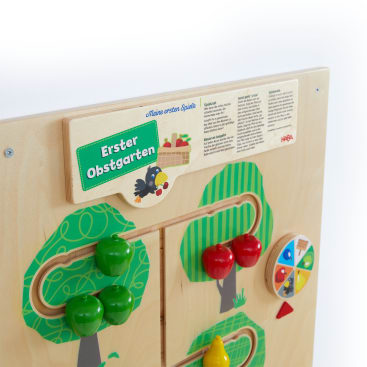 Ein hölzernes, an der Wand montiertes Spielbrett mit dem Namen Erster Obstgarten mit bunten Holzobstteilen, einem Spinnrad und bemalten Bäumen, auf dem die Kinder spielen und etwas über die Obsternte lernen können.