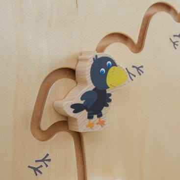 Ein Holzspielzeug mit einer schwarzen Vogelillustration ist an einer gebogenen Metallschiene auf einer hellen Holzplatte befestigt. Entlang der Schiene sind Fußabdrücke von Vögeln aufgedruckt.