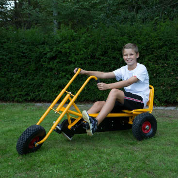 Ein Junge, der ein weißes T-Shirt, schwarze Shorts und Turnschuhe trägt, sitzt auf einem gelben Go-Kart mit drei Rädern auf einer Wiese mit Büschen und Bäumen im Hintergrund.