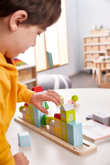 Ein Kind in einem gelben Kapuzenpulli ordnet bunte Holzbausteine in Form von Häusern und Bäumen auf einem Tisch in einem Klassenzimmer oder Spielzimmer an.