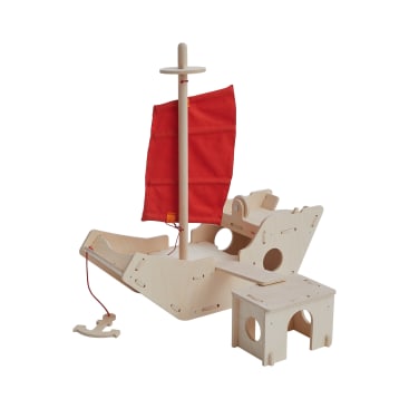 Ein Holzspielgerät in Form eines Bootes mit einem roten Stoffsegel, einem Steuerrad und einer kleinen Bank. Die Struktur ist aus leichtem Holz und hat ausgeschnittene Löcher und eine Zugschnur an der Vorderseite.