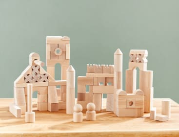 Eine Gruppe von Holzbausteinen, die auf einem Tisch so angeordnet sind, dass sie Türme, Bögen und geometrische Formen bilden, mit zwei kleinen Holzklötzchen davor. Der Hintergrund ist eine schlichte, hellgrüne Wand.