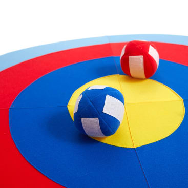 Nahaufnahme einer bunten Zielmatte, auf der ein roter und ein blauer weicher Ball liegen. Die Matte hat konzentrische Kreise in Rot, Blau und Gelb. Die Bälle haben weiße Flecken.