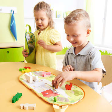 Zwei kleine Kinder spielen an einem Tisch mit tierförmigen Holzteilen und einer ausklappbaren Spielmatte. Der Junge legt ein Stück auf die Matte, während das Mädchen eine offene Tasche hält. Das Zimmer ist hell mit grünem und gelbem Dekor.