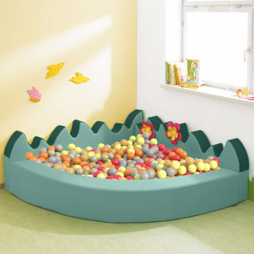 Ein grüner, mit Schaumstoff gepolsterter Ballkasten, der mit gelben, orangefarbenen, roten und grauen Plastikbällen gefüllt ist, steht in der Ecke eines Zimmers in der Nähe eines Fensters. Die Wand ist mit zwei ausgestanzten Vögeln und einer Blume dekoriert.