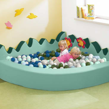 Zwei kleine Kinder liegen in einem hellgrünen, mit bunten Plastikbällen gefüllten Bällebad. Die Kugelgrube ist mit einem gewellten Rand und Blumenformen verziert und befindet sich in einem hellen Raum mit Wanddekorationen und Büchern.