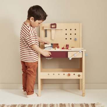 Ein kleines Kind in einem gestreiften Hemd und einer rostfarbenen Hose steht an einer hölzernen Spielwerkbank und spielt mit Spielzeugwerkzeugen und Zubehör. Die Werkbank steht vor einer beigen Wand auf einem gemusterten Teppich.
