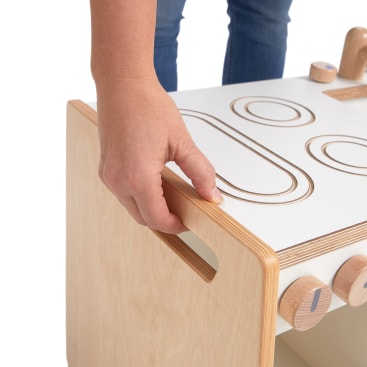 Eine Person ergreift den Griff eines weißen Aktivitätswürfels aus hellem Holz mit Rillen und Knöpfen, während sie auf einem weißen Hintergrund steht.