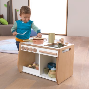 Ein kleines Kind steht an einem hölzernen Spielküchen-Set mit Spielzeugtöpfen, -pfannen und -geschirr und scheint mit einem Löffel etwas umzurühren. Die Szene spielt in einem Innenraum auf einem Holzboden mit weichen Möbeln im Hintergrund.