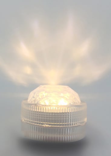 Eine kleine, runde, durchsichtige Kunststoff-LED-Leuchte sitzt auf einer glatten Oberfläche, strahlt ein helles, weißes Licht nach oben ab und erzeugt radiale Muster auf dem Hintergrund.