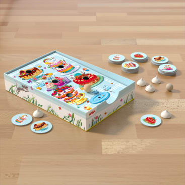 Eine Brettspielschachtel für Kinder mit Cartoon-Desserts und Insekten auf dem Deckel steht auf einem Holzboden. Runde, illustrierte Spielsteine und weiße, kegelförmige Spielfiguren sind neben der Schachtel angeordnet.