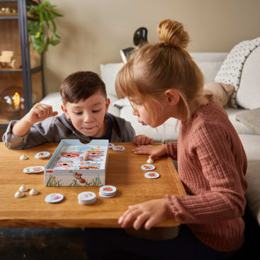 Zwei kleine Kinder sitzen an einem Holztisch und spielen konzentriert ein Brettspiel mit runden Figuren. Spielfiguren und Karten sind ausgebreitet, und im Hintergrund ist eine Couch mit Kissen zu sehen.