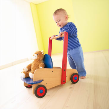 Ein Kleinkind in blauer Kleidung schiebt einen hölzernen Lauflernwagen mit roten Griffen und Rädern. Ein brauner Teddybär sitzt auf dem blauen Sitz der Gehhilfe in einem hell erleuchteten Raum mit hellem Holzboden und gelben Wänden.